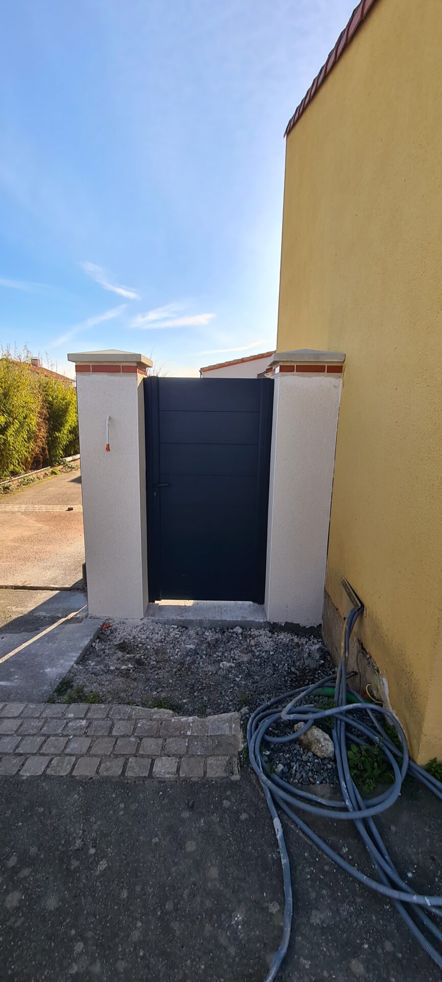 Portillon entre deux piliers en béton à coté d'une maison aux murs jaunes